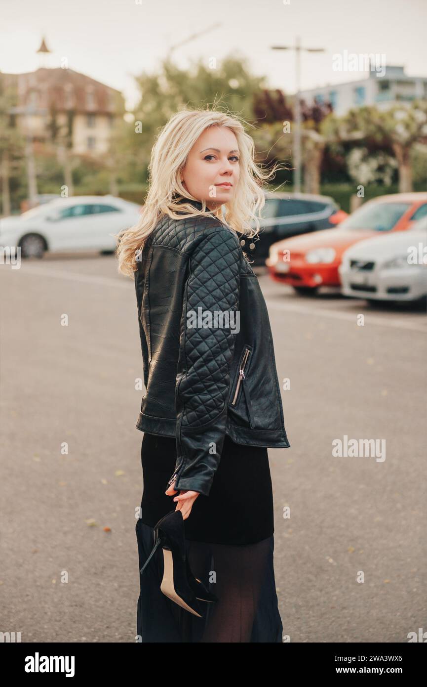 Außenporträt einer schönen blonden Frau, die schwarze Lederjacke trägt, hohe Schuhe hält und weggeht Stockfoto