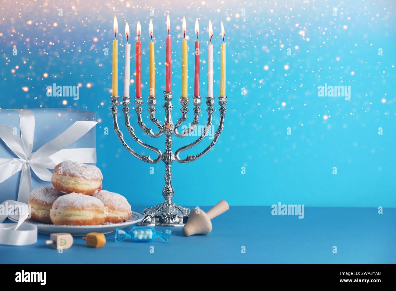 Hanukkah-Feier. Menora mit brennenden Kerzen, Dreidels, Donuts und Geschenkbox auf hellblauem Tisch, Platz für Text Stockfoto