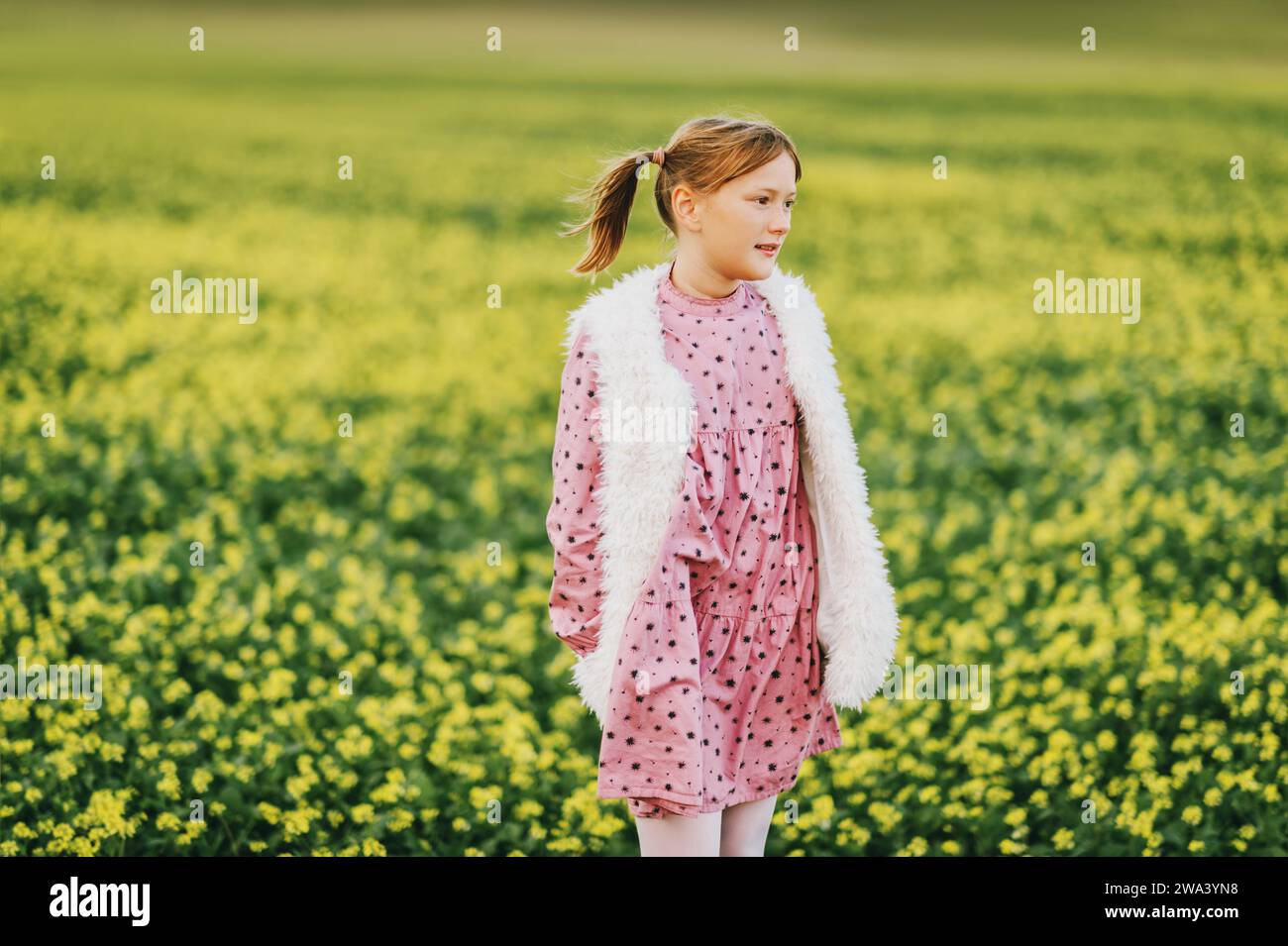Außenporträt eines hübschen 10-jährigen Mädchens mit rosa Vintage-Kleid und Kunstfell-Bodywärmer, der auf einem Feld posiert Stockfoto