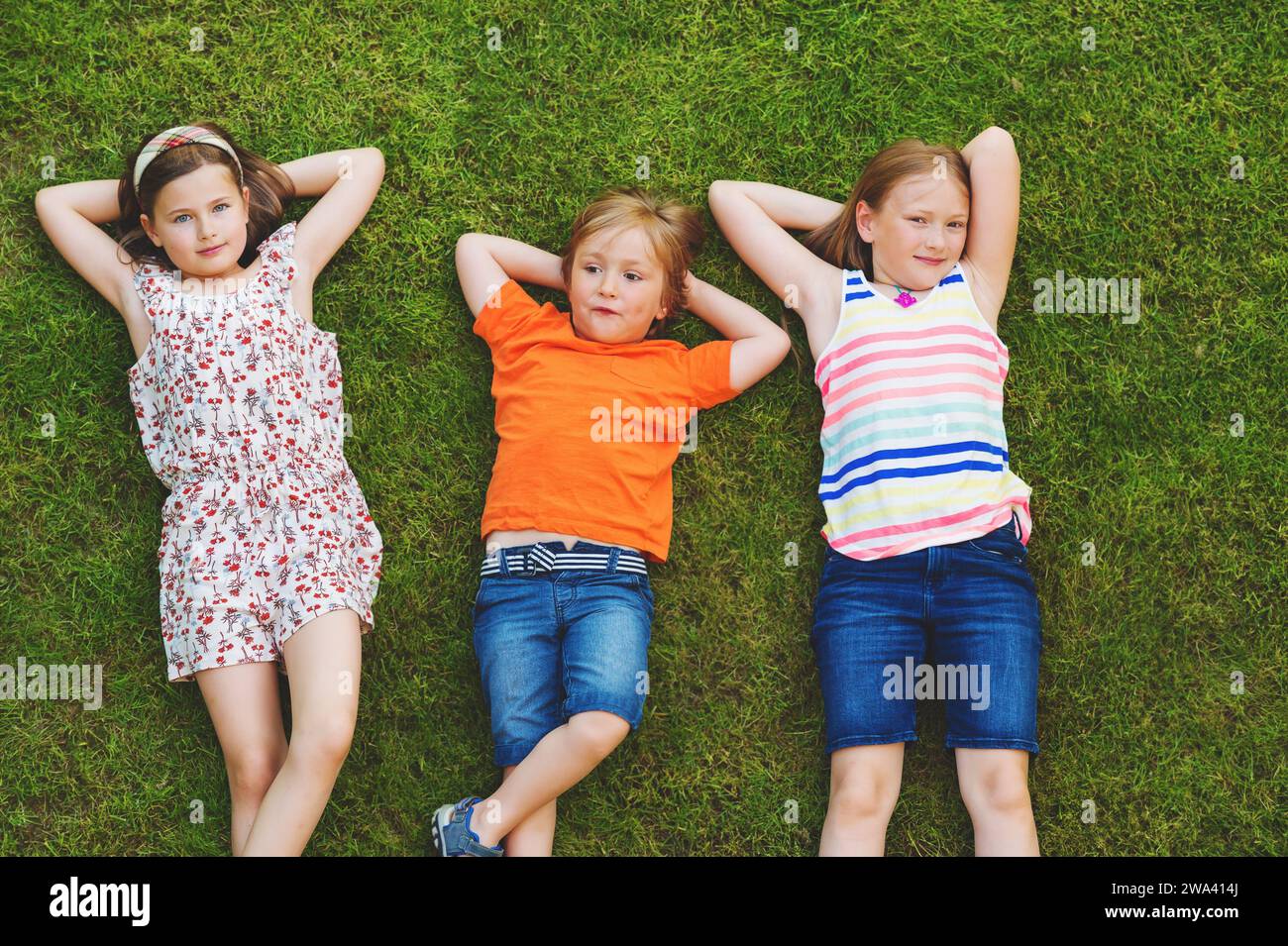 Glückliche Kinder, die sich im Freien amüsieren. Kinder spielen im Sommerpark. Kleiner Junge und zwei Mädchen liegen auf grünem, frischem Gras Stockfoto