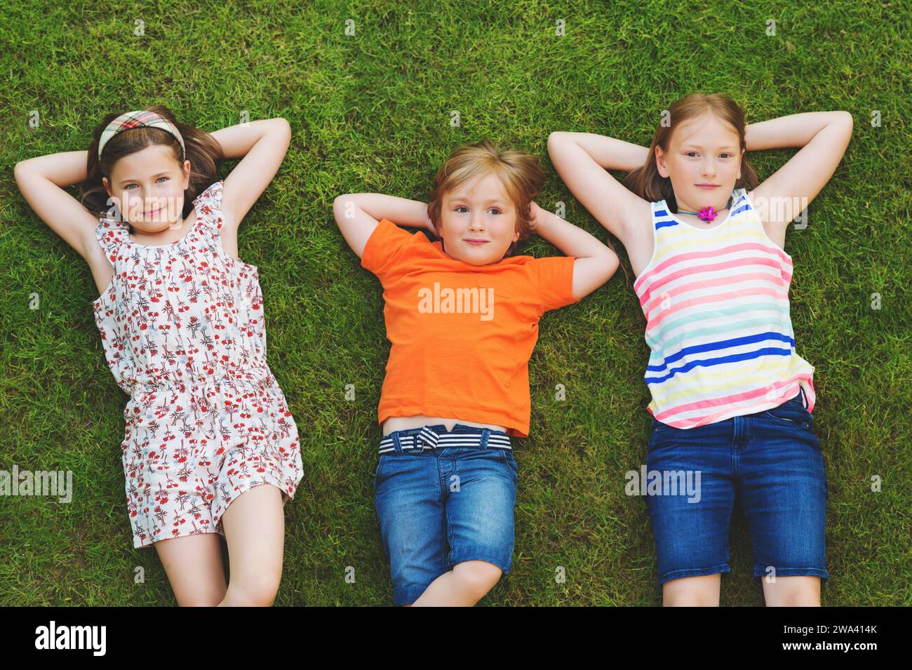 Glückliche Kinder, die sich im Freien amüsieren. Kinder spielen im Sommerpark. Kleiner Junge und zwei Mädchen liegen auf grünem, frischem Gras Stockfoto