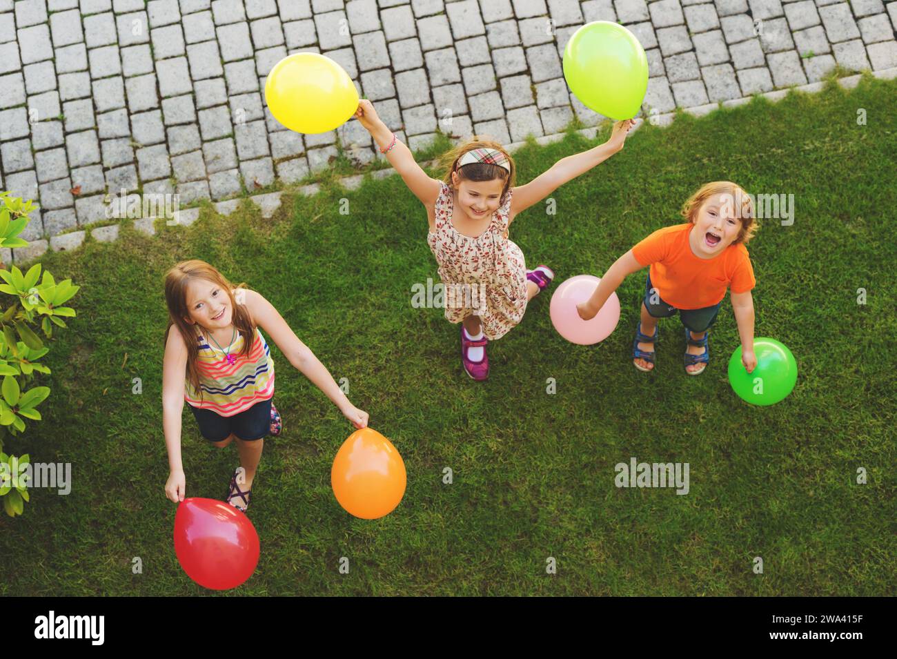 Drei fröhliche kleine Kinder spielen mit bunten Ballons draußen, Draufsicht Stockfoto