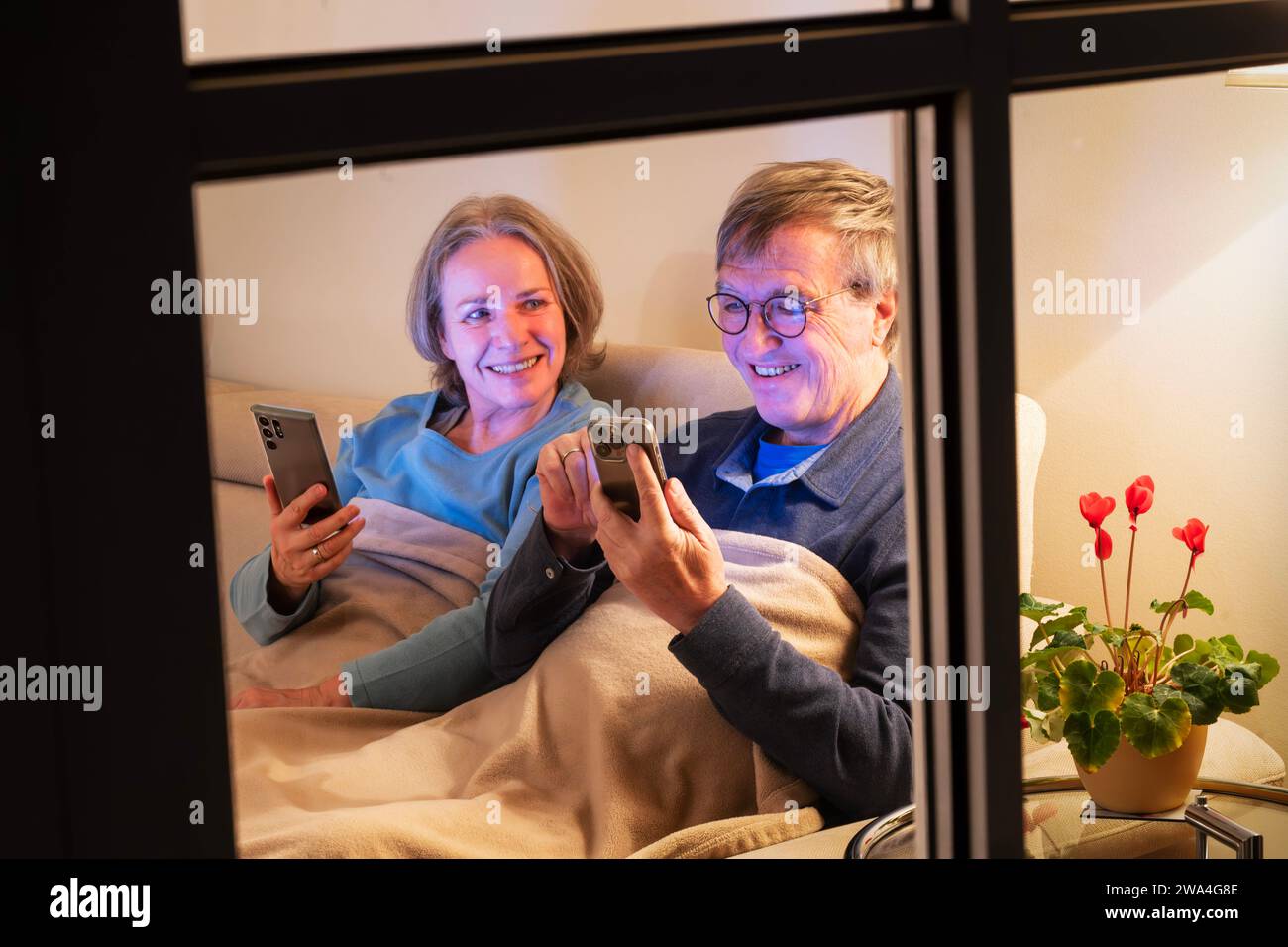 Seniorenpaar surft abends im Internet, beide sitzen mit ihren Smartphones gemütlich auf dem Sofa, München, Januar 2024 Deutschland, München, Januar 2024, Paar sitzt abends gemütlich zuhause, sie surfen mit ihren Handys im Internet, lachen, reden, haben Spaß miteinander, das Paar, Ü60, sitzt gemeinsam auf dem Sofa, gemütlich in eine Decke eingekuschelt, jeder mit seinem eigenen Smartphone, Handys von Samsung und Apple, bequem, gemütlich, hyggelig, beide über sechzig, Babyboomer, Leben im Alter, Rentner, Partnerschaft, Wohnzimmer, auf dem Beistelltisch stehen ein Alpenveilchen, Blick von draussen Stockfoto