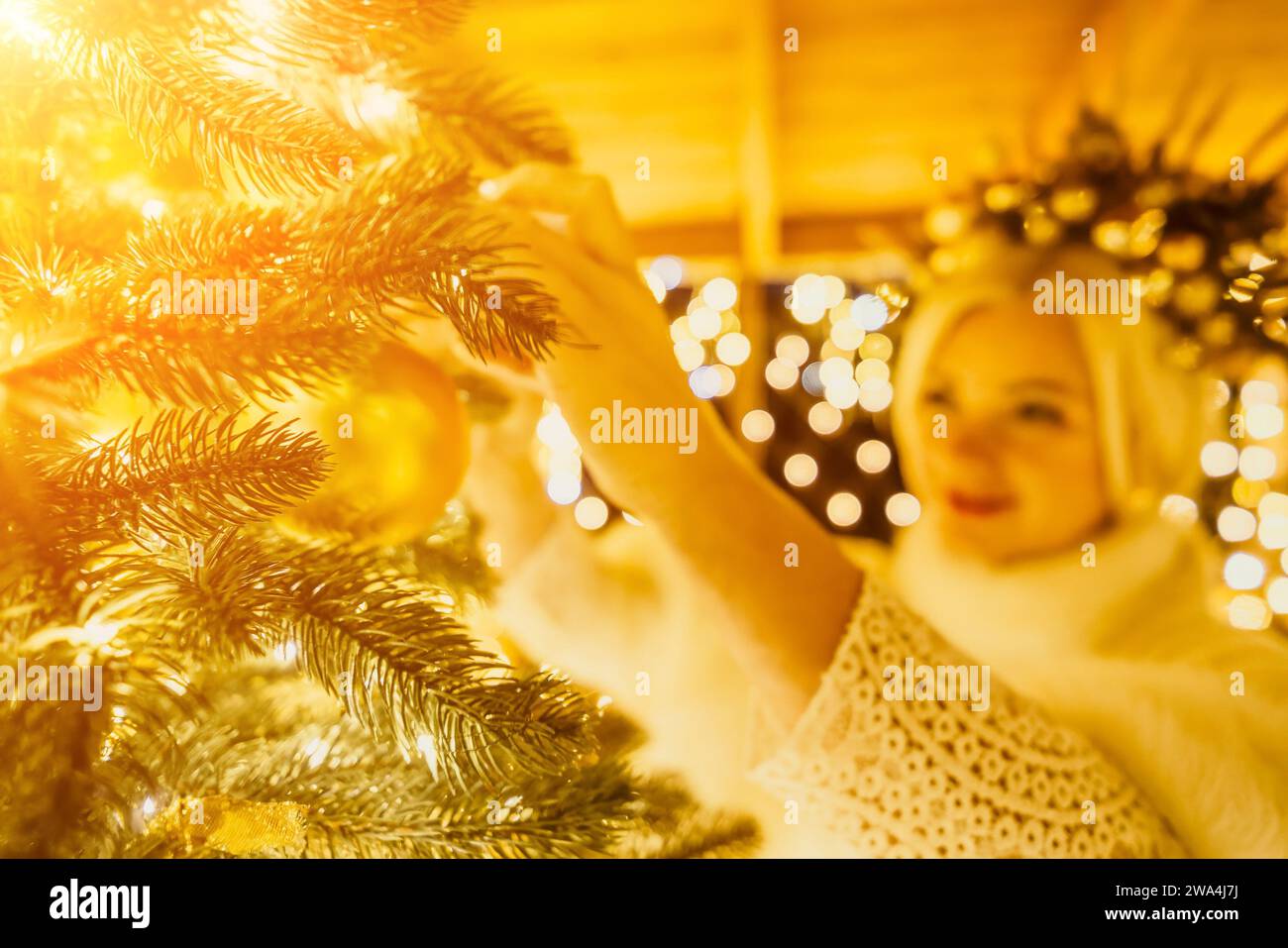 Eine blonde Frau in weißem Kleid und eine Krone aus goldenen Ornamenten schmücken den Weihnachtsbaum mit goldenen Ornamenten und Lichtern. Der Baum ist mit Gold verziert Stockfoto