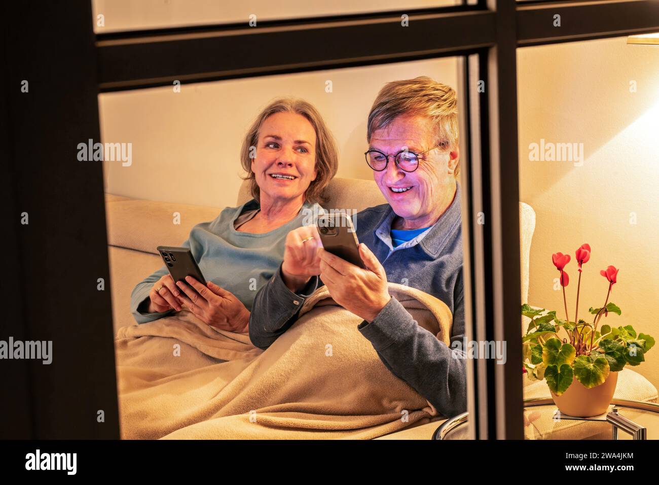 Seniorenpaar surft abends im Internet, beide sitzen mit ihren Smartphones gemütlich auf dem Sofa, München, Januar 2024 Deutschland, München, Januar 2024, Senioren lesen abends in ihren Handys, das Paar, Ü60, sitzt zuhause gemeinsam auf dem Sofa, gemütlich in einer Decke eingekuschelt, jeder schaut in sein eigenes Smartphone, Handys von Samsung und Apple, die beiden Babyboomer surfen im Internet, bequem, gemütlich, hyggelig, beide über sechzig, Leben im Alter, Rentner, Partnerschaft, Wohnzimmer, auf dem Beistelltisch steht ein Alpenveilchen, Blick von draussen durchs Fenster, Ehepaar, Gemeinsamk Stockfoto