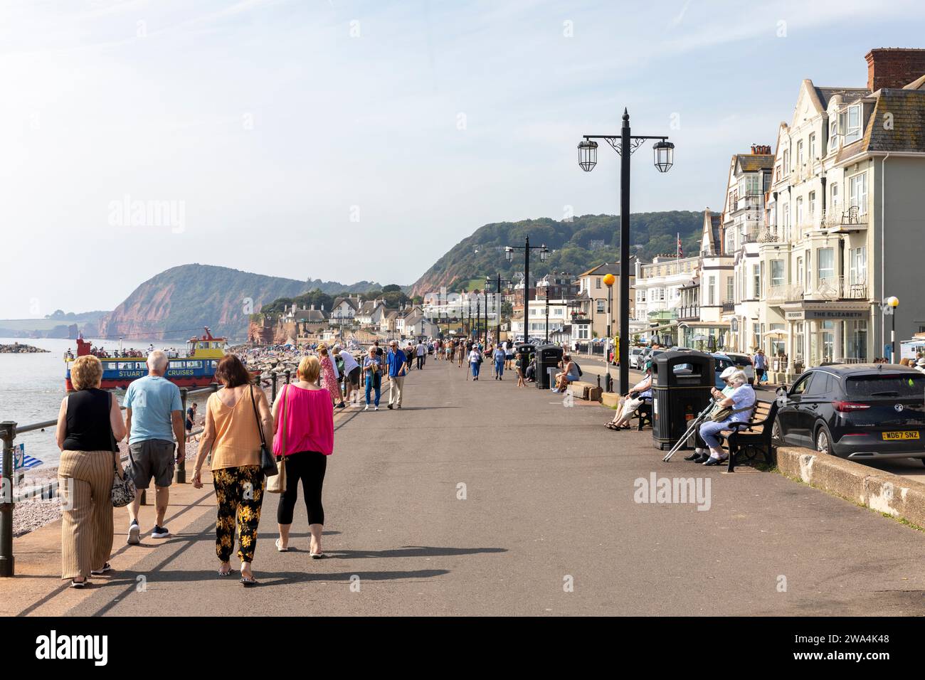 Sidmouth Dorset 2023, die Leute laufen entlang der Sidmouth Promenade am Meer mit jurastude im Hintergrund, England, Großbritannien Stockfoto