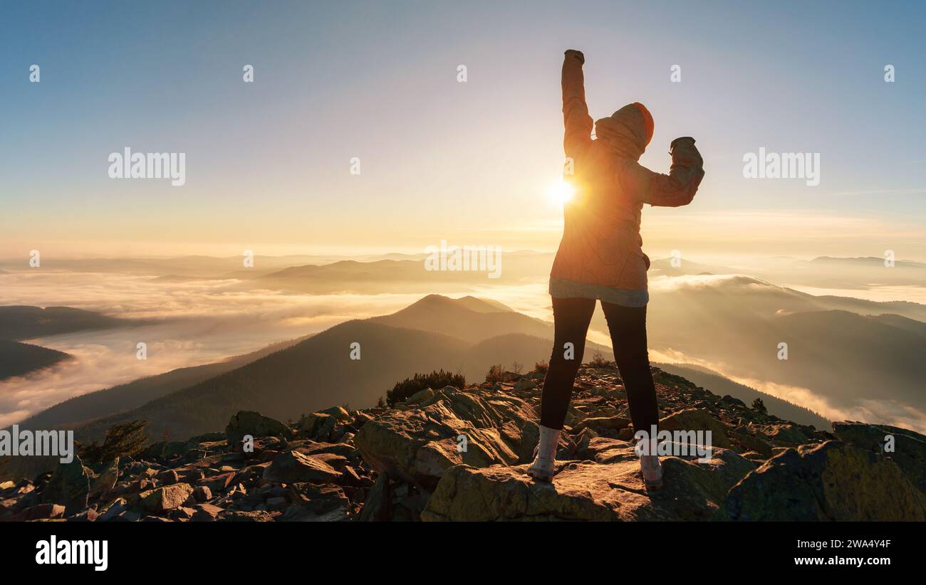 Erfolgreiches Wandermädchen, das mit erhobenen Armen auf dem Berggipfel steht. Mädchen begrüßt die Sonne bei Sonnenaufgang. Leistungskonzept. Stockfoto