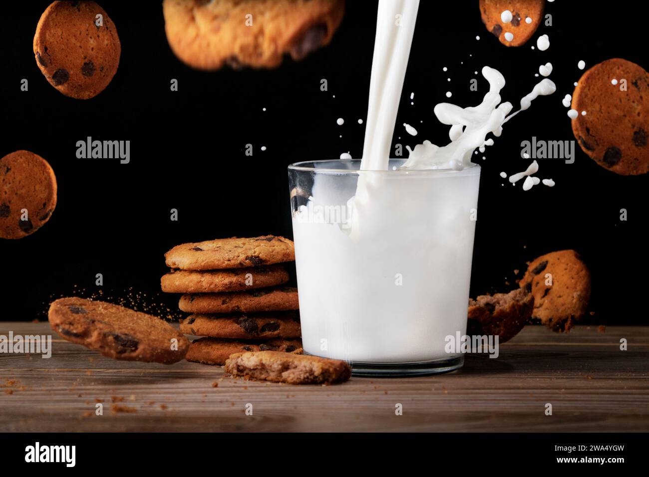 Ein Glas Milch mit leckeren Keksen. Milch wird mit Spritzern in eine Tasse gegeben. Kekse fliegen in der Luft und fallen mit Krümeln auf den Tisch. Schwarzer Hintergrund Stockfoto