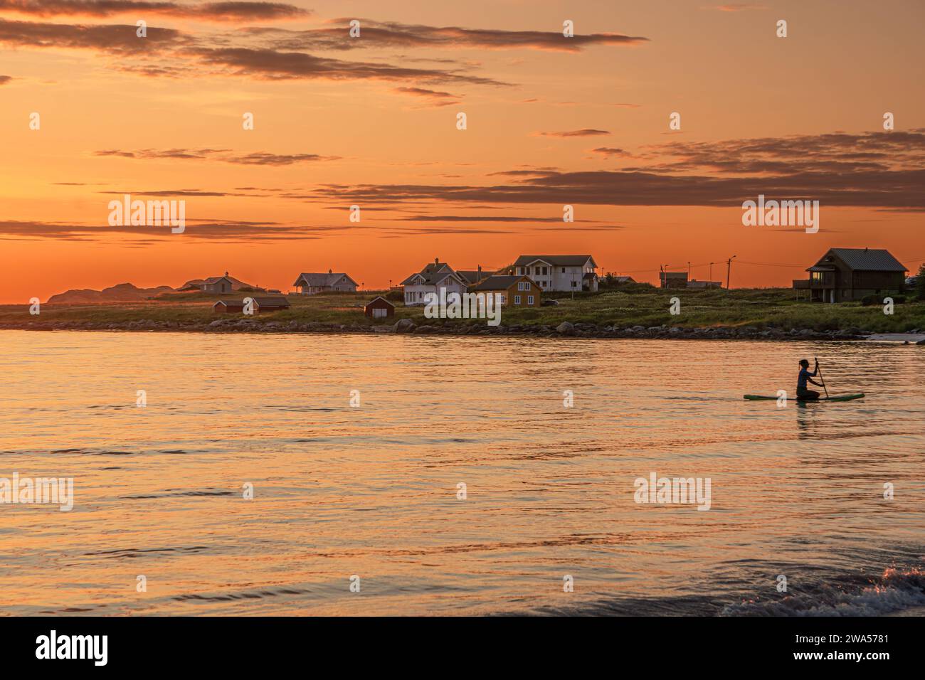 Eine Frau im Kajak in der Nähe von Ramberg, Lofoten, Norwegen, während einer warmen Mittsommernacht, Häuser im Hintergrund, orange Sonnenuntergang, teilweise bewölkt. Stockfoto
