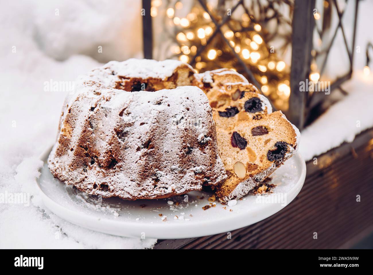Traditioneller Weihnachtskuchen oder Obstkuchen mit getrockneten Früchten auf Teller mit ausgeschnittenen Stücken, Schnee und Weihnachtsbeleuchtung auf Hintergrund. Stockfoto