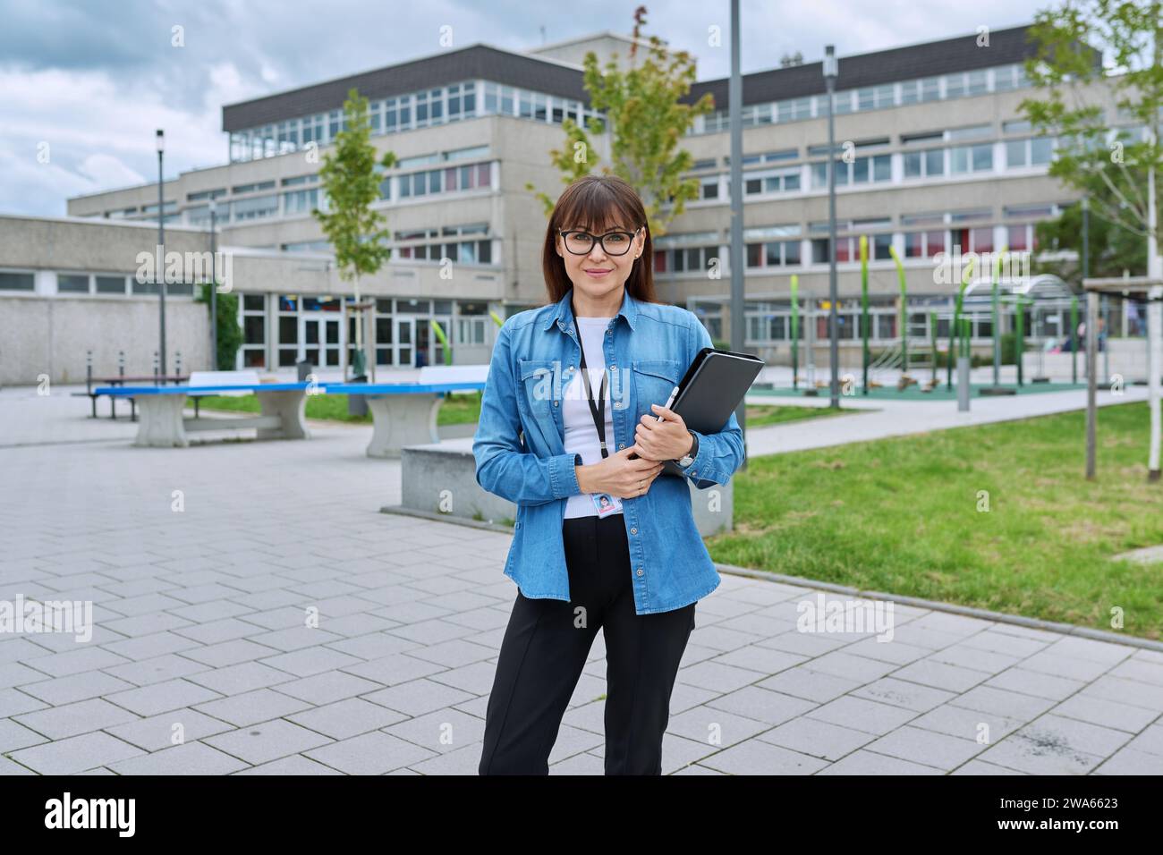 Schullehrerin mittleren Alters mit digitalem Tablet posiert in der Nähe des Schulgebäudes Stockfoto