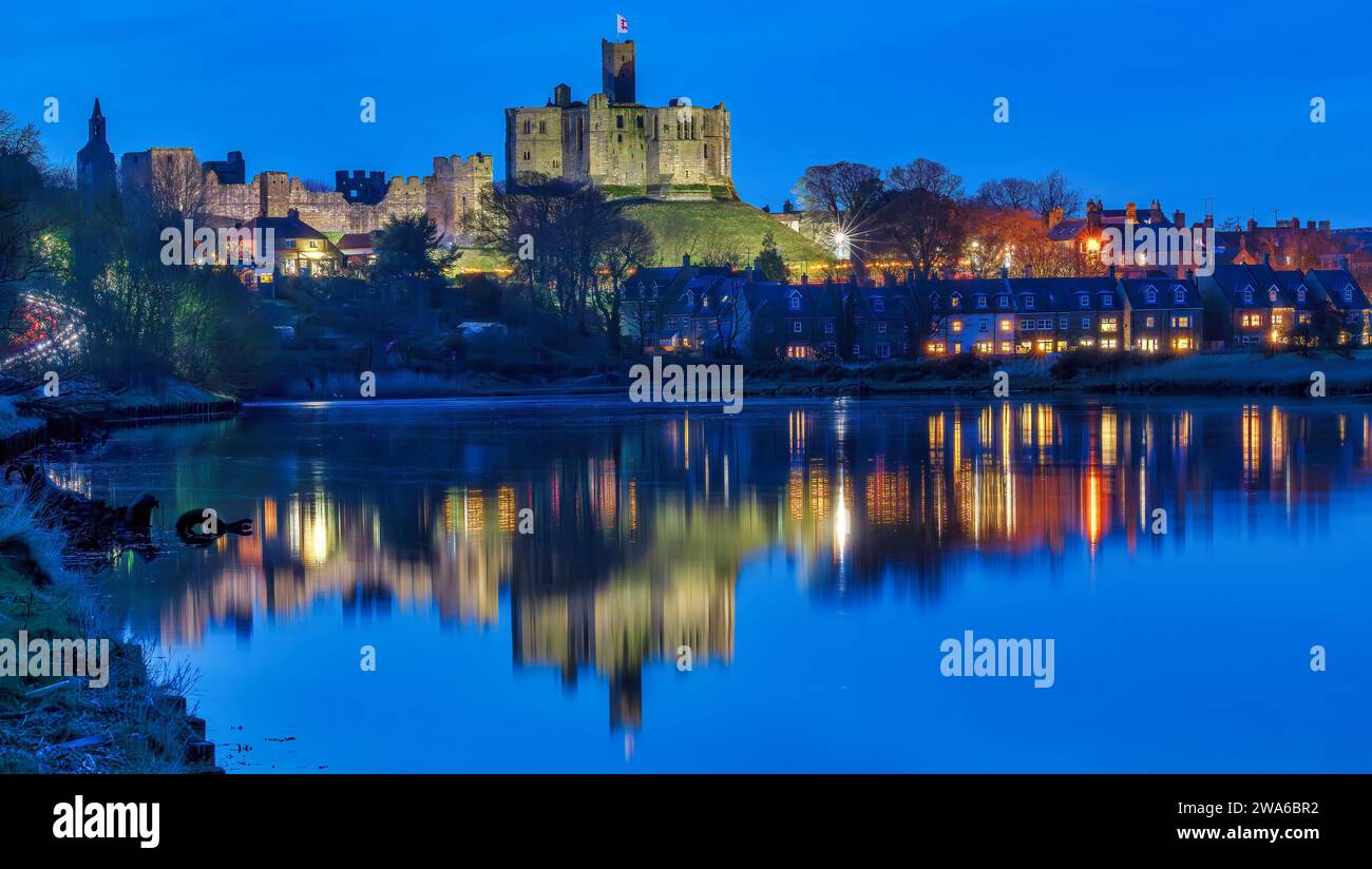 Blick in der Abenddämmerung auf Warkworth Castle und Warkworth Village, die sich im Fluss Coquet spiegeln, wie man von einem Spaziergang mit Warkworth Castle im Flutlicht sehen kann. Stockfoto