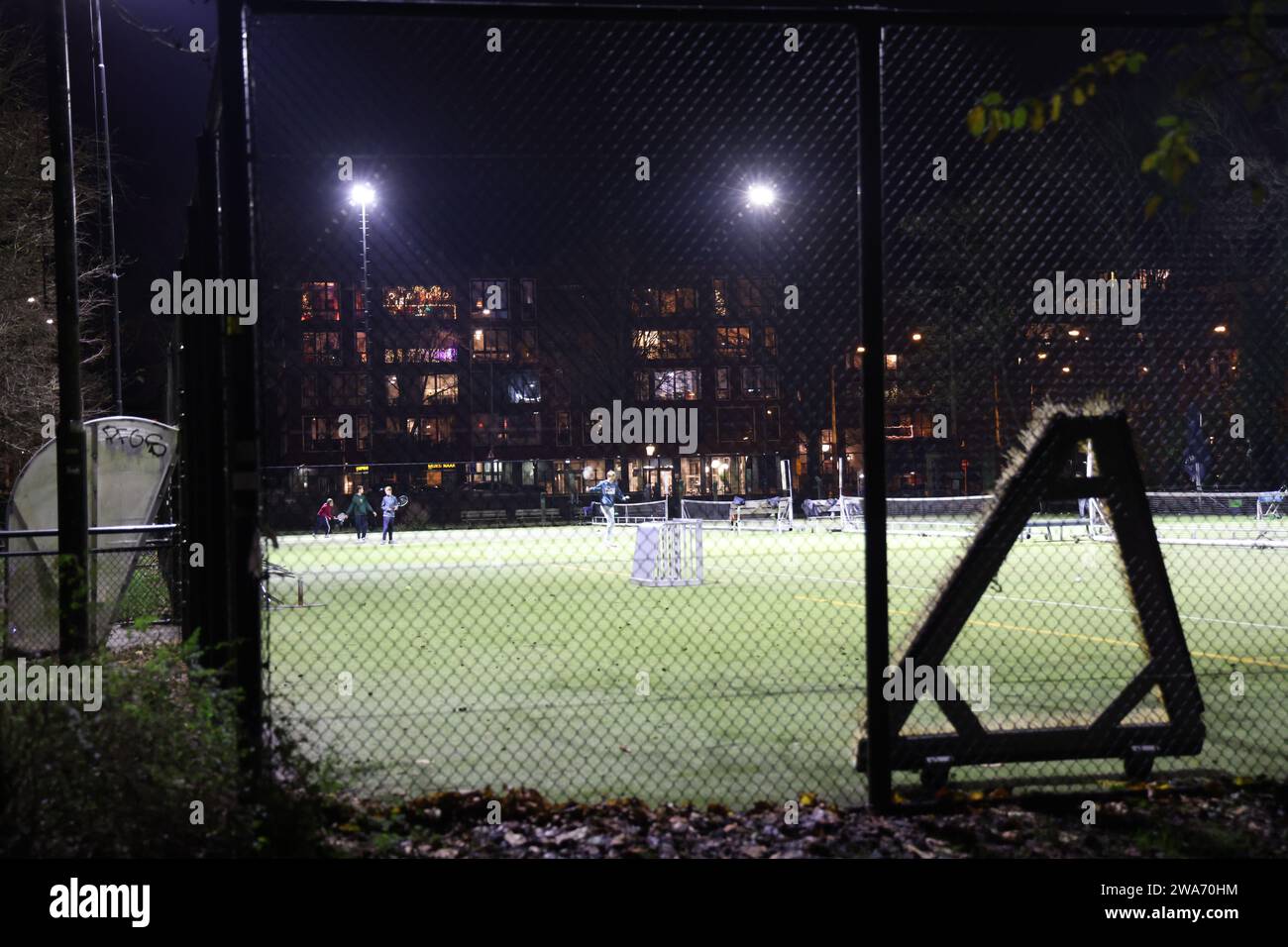 Sportler spielen abends Tennis auf einem beleuchteten Feld in der Dunkelheit, durch einen Zaun gesehen Stockfoto