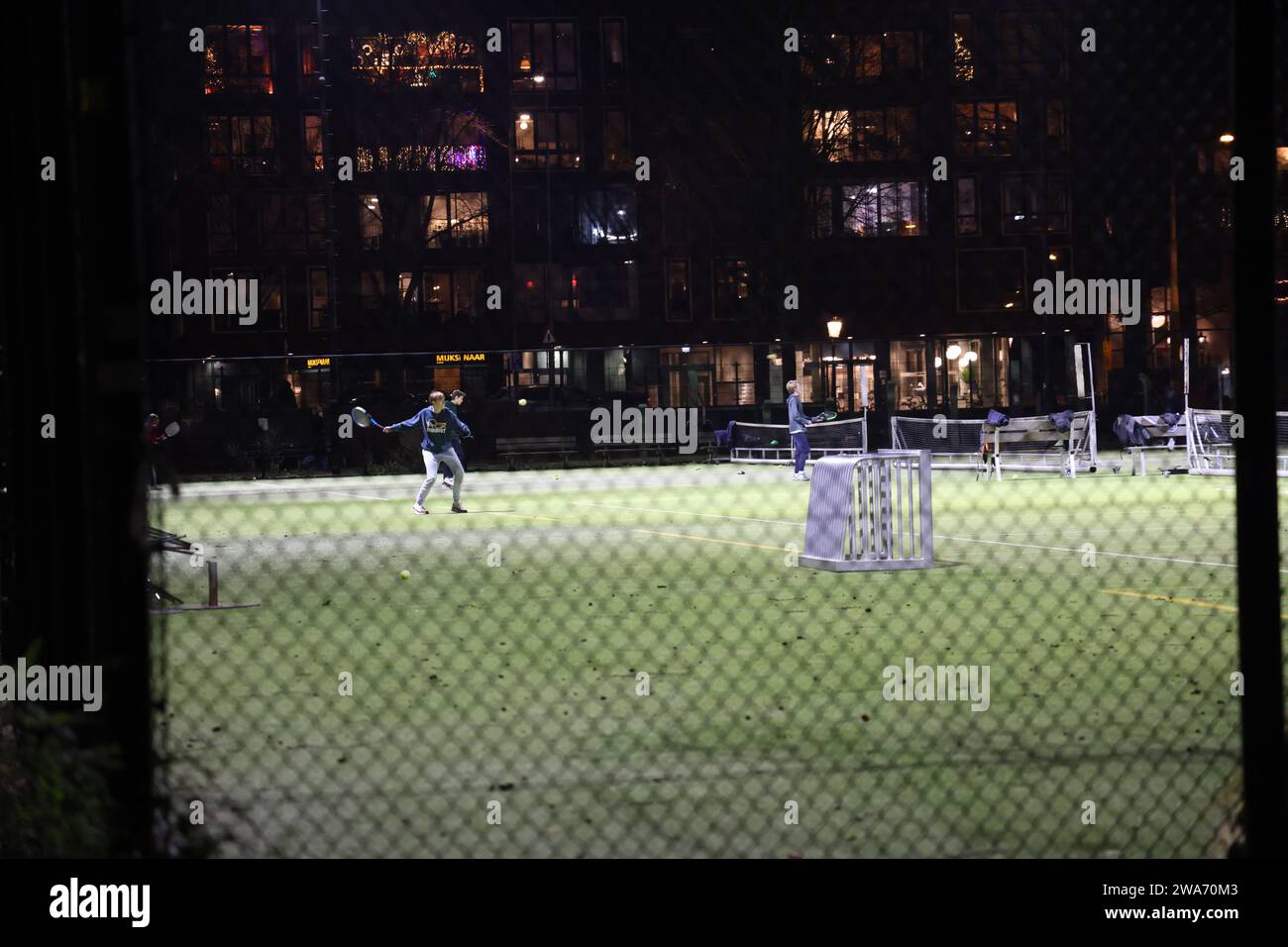 Sportler spielen abends Tennis auf einem beleuchteten Feld in der Dunkelheit, durch einen Zaun gesehen Stockfoto