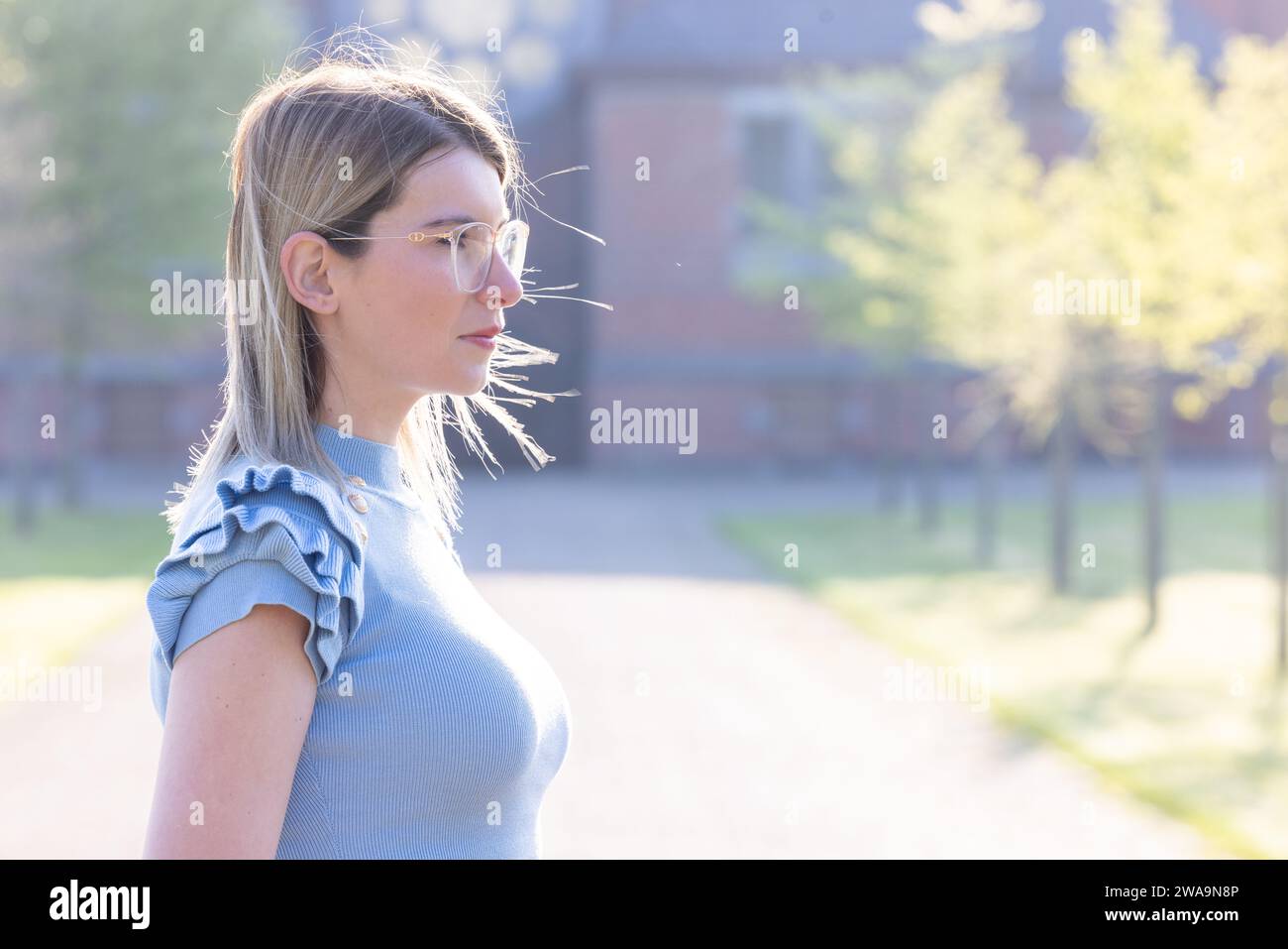 Ein Seitenprofil einer jungen kaukasischen Frau mit Brille und einem blauen Oberteil mit Rüschendetails steht vor einem weichen Hintergrund einer üppigen Frühlingseinstellung. Ihr Haar ist ordentlich zurückgebunden, und das Sonnenlicht hebt sanft ihre Gesichtszüge und die Haarkanten hervor. Der Hintergrund deutet auf einen ruhigen Park- oder Gartenweg hin, der zum friedlichen und reflektierenden Thema des Bildes beiträgt. Frühjahrstagsbesinnung. Hochwertige Fotos Stockfoto