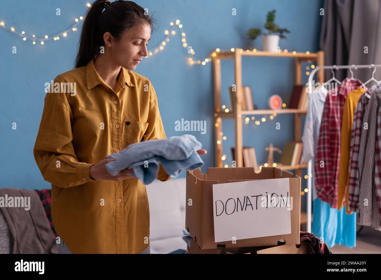 Die hispanische Frau sammelt zu Hause Kleidung in einer Spendenbox. Spendenbox für Arme mit Kleidung im Wohnzimmer. Lady spendet Kleidung für den Unterschlupf. Stockfoto