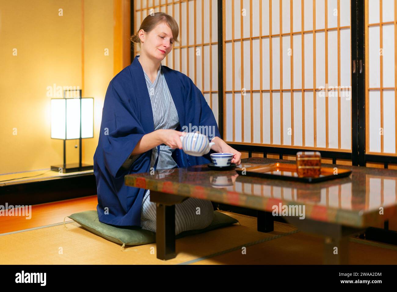Eine junge kaukasische Frau in japanischer Kleidung sitzt am Tisch im Ryokan-Raum und gießt Tee aus Stockfoto