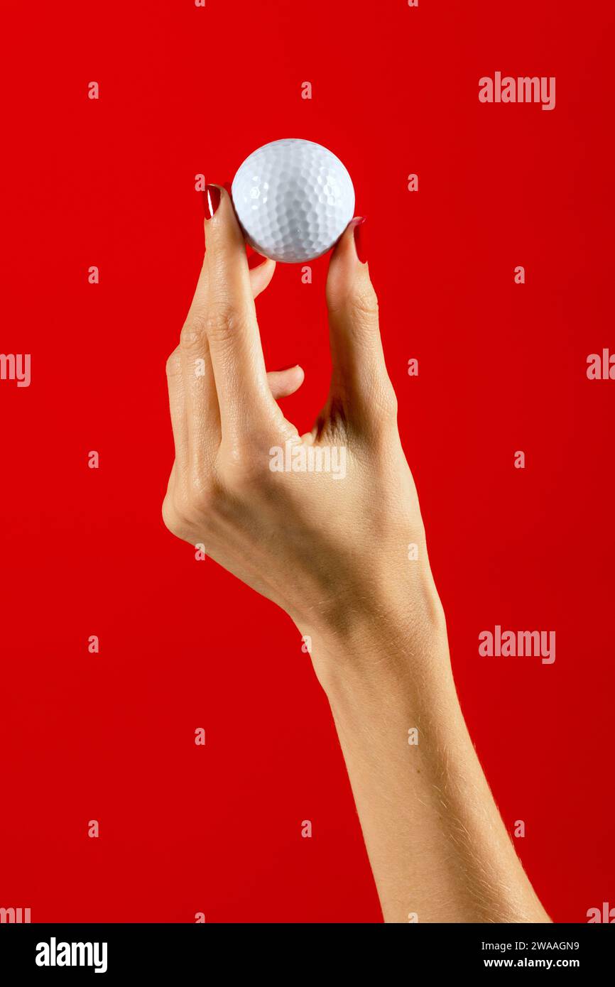 Beschneiden Sie anonyme Frau mit Maniküre, die kleinen weißen Golfball vor rotem Hintergrund hält. Foto des Golfkonzepts. Stockfoto