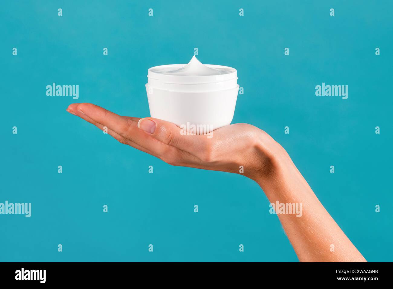 Anonyme junge schlanke weibliche Hand, die ein weißes Glas mit Körpercreme in der Hand zeigt, mit gepflegten Fingern in Licht vor türkisfarbenem Hintergrund Stockfoto