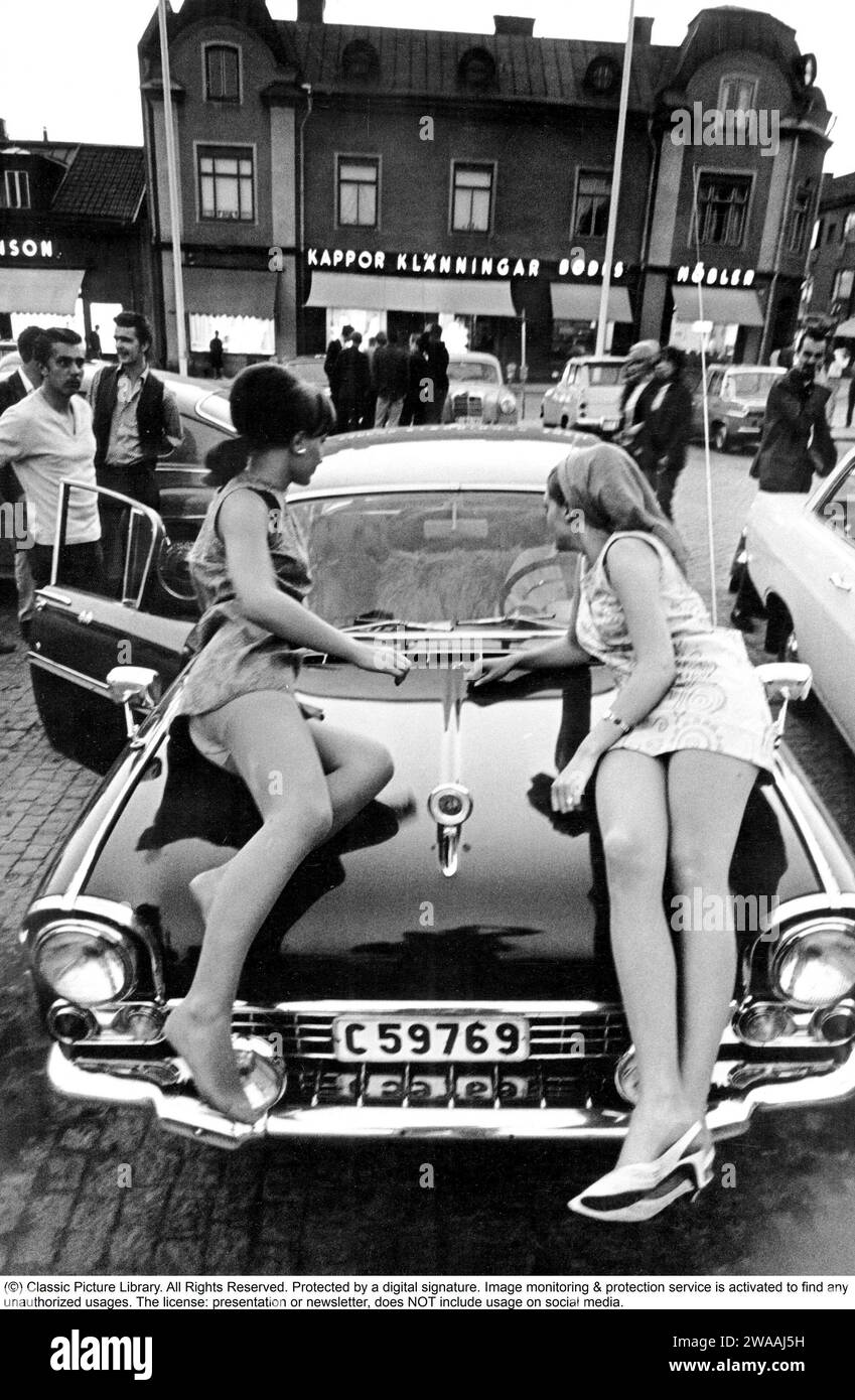In den 1960er Jahren Zwei Mädchen liegen auf der Motorhaube eines amerikanischen Oldtimers, der auf einem Platz in einer kleinen Stadt in Schweden geparkt ist. Eine typische Jugendmotorkultur aus den 1960er Jahren, in der oft nachts gemeinsam restaurieren, fahren und Spaß haben. Schweden 1964 Stockfoto