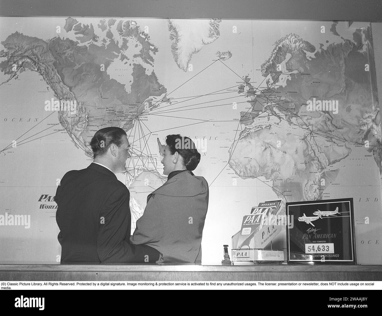 In den 1950er Jahren Ein junges Paar in einem Reisebüro steht vor einer Weltkarte und betrachtet alle Flugrouten der amerikanischen Fluggesellschaft Pan American zwischen verschiedenen Ländern und Kontinenten. Das klassische Logo von Pan am ist auf der Karte und auf dem Ordner auf dem Tresen zu sehen. Die Fluggesellschaft war die größte internationale Fluggesellschaft in den USA mit den meisten internationalen Flügen. 1955. Kristoffersson Ref. BX51-4 Stockfoto
