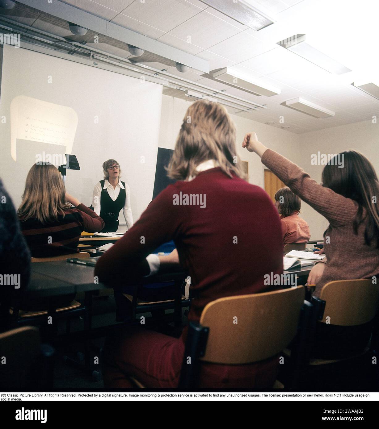 In den 1970er Jahren Eine Schulklasse sitzt während eines Unterrichts mit einer Lehrerin auf den Bänken. Vorne steht ein Overheadprojektor, der damals in Klassenzimmern üblich war und das transparente Blatt mit Text und Bildern auf dem Gerät auf dem Bildschirm zeigte. Overheadprojektoren wurden vor der Einführung computergestützter Projektionen in Bildung und Wirtschaft weit verbreitet. Schweden 1970er Jahre Stockfoto