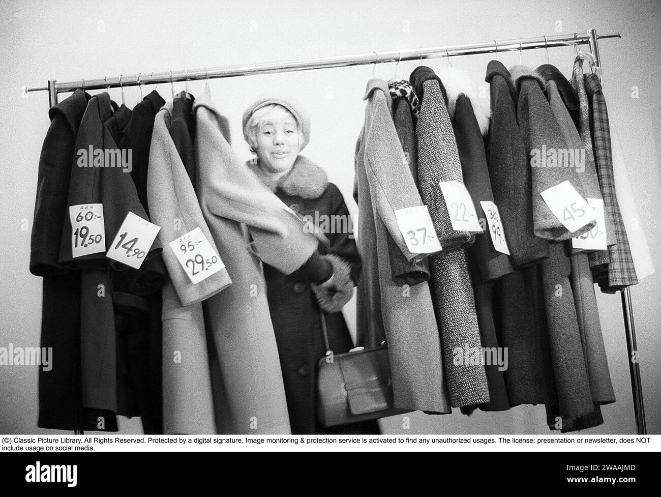 Tolle Rabatte auf Oberbekleidung in den 1960ern Eine Frau steht an einem Kleiderständer und wählt aus Oberbekleidung und Mänteln, die mit Preisschildern mit ermäßigten Preisen gekennzeichnet sind. Nach ihrem Gesicht zu urteilen, schätzt sie die reduzierten Preise und denkt, sie bekommt echte Schnäppchen. Schweden Januar 1963. Ref. CV28 Stockfoto
