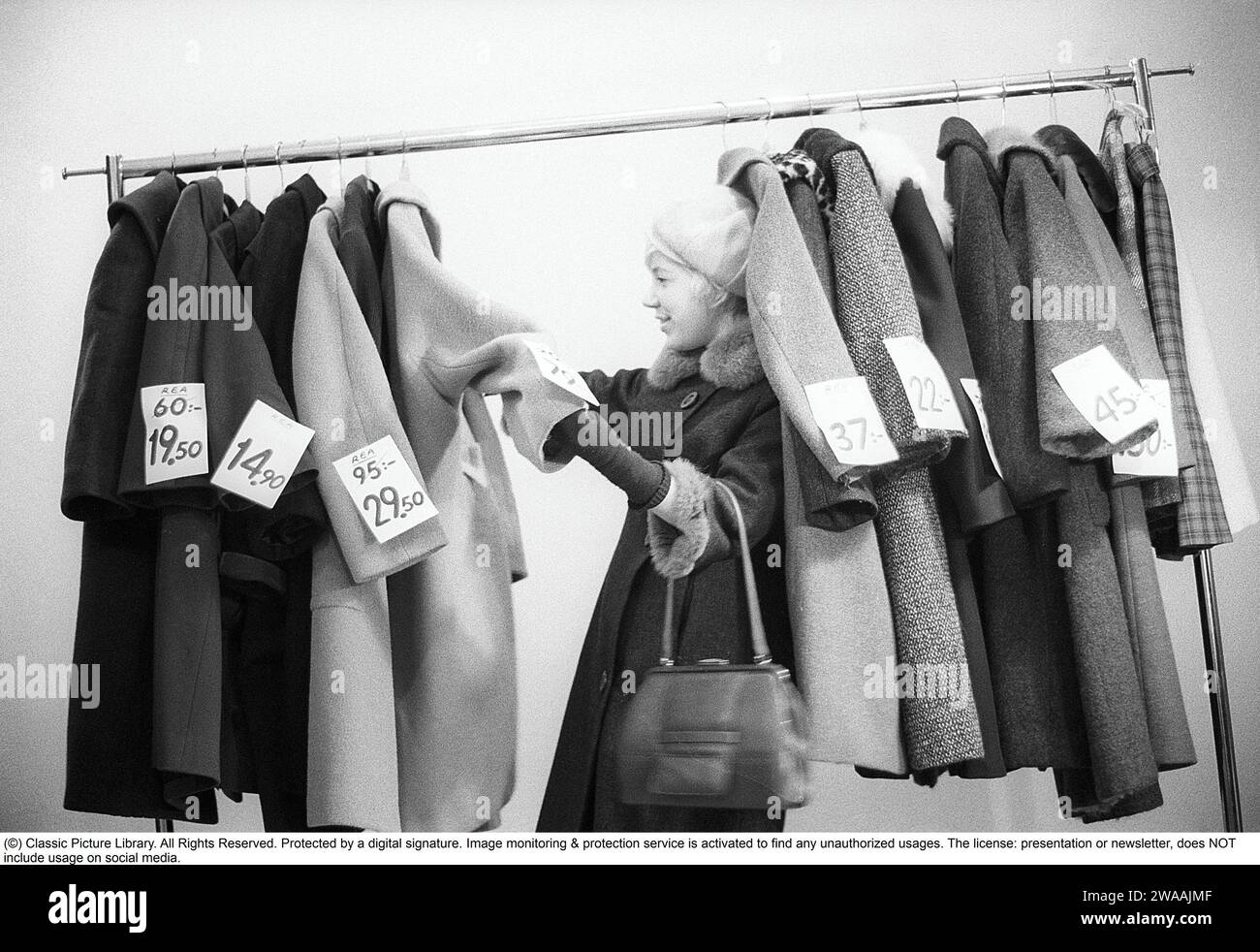 Tolle Rabatte auf Oberbekleidung in den 1960ern Eine Frau steht an einem Kleiderständer und wählt aus Oberbekleidung und Mänteln, die mit Preisschildern mit ermäßigten Preisen gekennzeichnet sind. Nach ihrem Gesicht zu urteilen, schätzt sie die reduzierten Preise und denkt, sie bekommt echte Schnäppchen. Schweden Januar 1963. Ref. CV28 Stockfoto