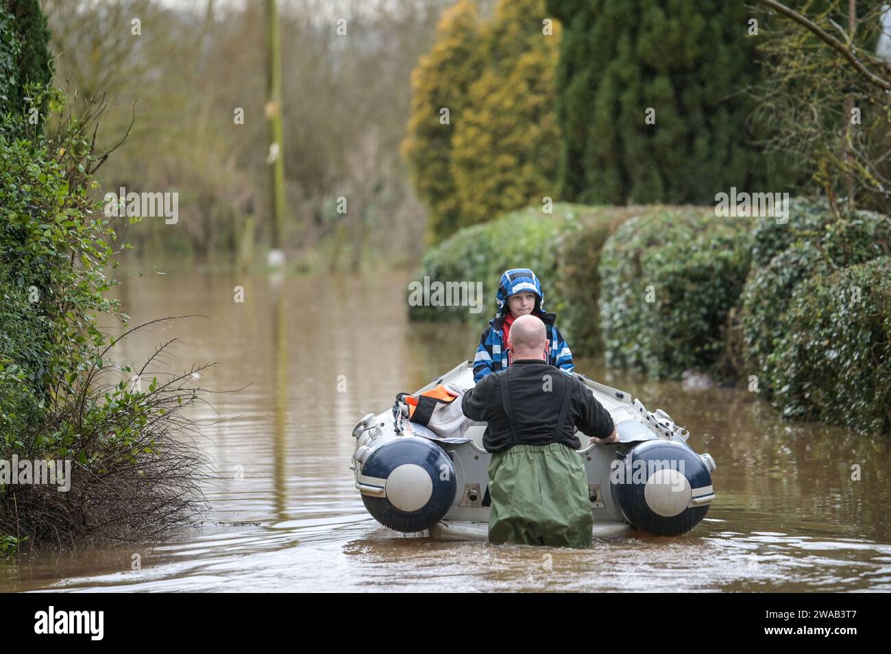 Worcester, 3. Januar 2024: Jack, siebeinhalb Jahre alt, wird mit dem Boot zu seinem Haus im englischen Dorf Severn Stoke gebracht, das durch Überschwemmungen vollständig abgeschnitten wurde, nachdem der Fluss Severn aufgrund anhaltender Regenfälle durch Sturm Henk seine Ufer geplatzt hatte. Der Rose and Crown Pub konnte gesehen werden, wie er Wasser herauspumpte, als er mit seinen selbstgemachten Hochwasserschutzanlagen um trocken kämpfte. - Credit: Stop Press Media/Alamy Live News Stockfoto