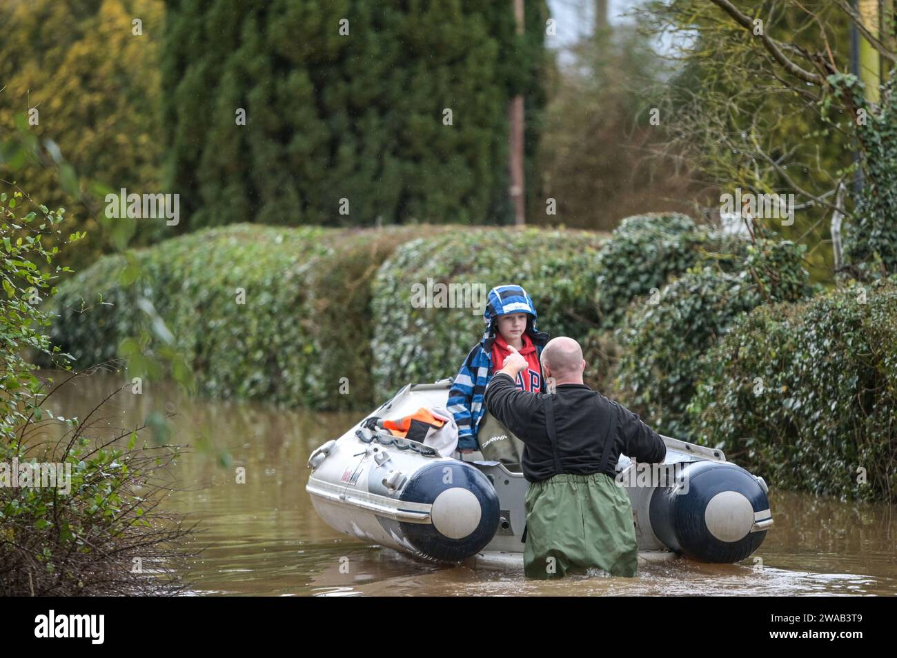 Worcester, 3. Januar 2024: Jack, siebeinhalb Jahre alt, wird mit dem Boot zu seinem Haus im englischen Dorf Severn Stoke gebracht, das durch Überschwemmungen vollständig abgeschnitten wurde, nachdem der Fluss Severn aufgrund anhaltender Regenfälle durch Sturm Henk seine Ufer geplatzt hatte. Der Rose and Crown Pub konnte gesehen werden, wie er Wasser herauspumpte, als er mit seinen selbstgemachten Hochwasserschutzanlagen um trocken kämpfte. - Credit: Stop Press Media/Alamy Live News Stockfoto