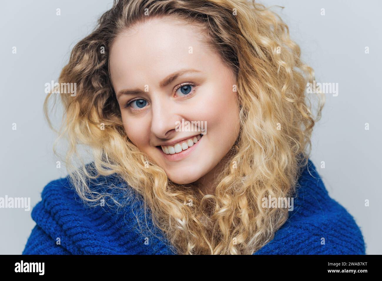 Fröhliche junge Frau mit leuchtend lockigen blonden Haaren, strahlendem Lächeln und einem kuscheligen blauen Strickpullover. Stockfoto