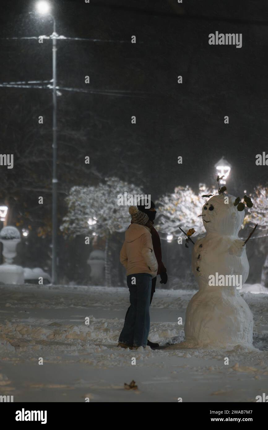 Ein Paar, das an einem verschneiten Abend handgemachte Schneemänner bewundert, die Parklichter glitzern Stockfoto