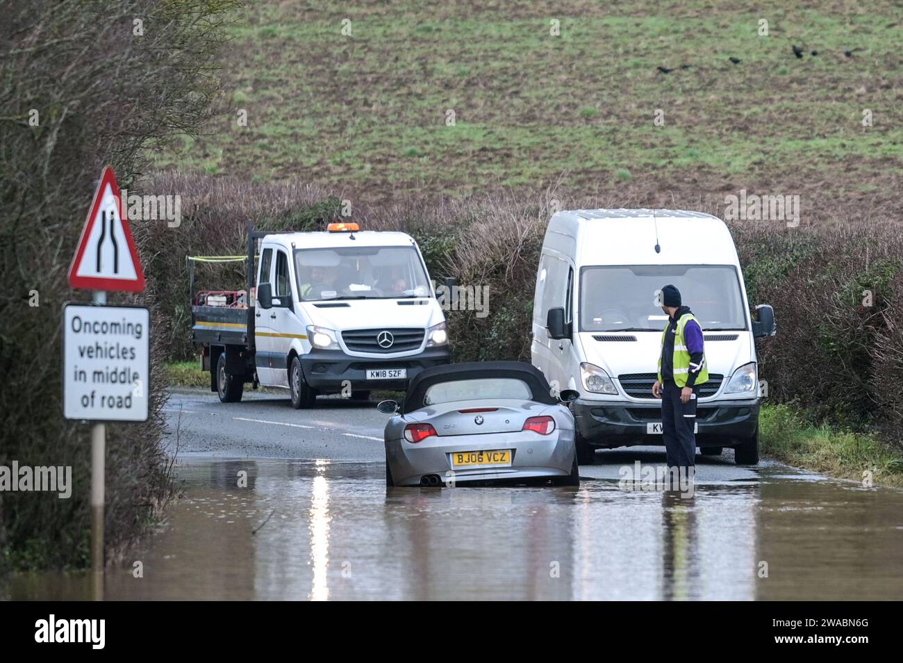 Offenham, Evesham, 3. Januar 2024 – Ein BMW-Fahrer versuchte und scheiterte, durch Hochwasser zu fahren, obwohl ein anderes Fahrzeug bereits auf derselben Strecke verlassen wurde. Der ältere Fahrer drehte seinen Motor auf, als er durchfuhr, aber seine Geschwindigkeit war zu hoch und Wasser stieg in seinen Motor ein. Das Fahrzeug ging schließlich wieder los, aber es kam nur wenige Meter vom Trockenland entfernt zu einer weiteren Haltestelle. Ein Straßenarbeiter kam dann an und legte als letzte Beleidigung ein Schild mit der Aufschrift „Road Closed“ vor sein Fahrzeug. Es geschah neben dem Offenham Caravan Park, der sich in der Nähe von Evesham in Worcestershire befindet, wo der Fluss Avon niederfließt Stockfoto