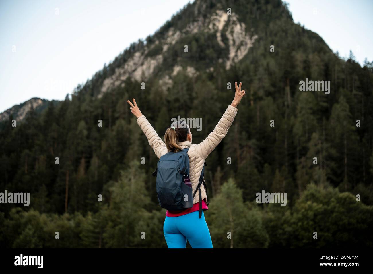 Junge Wanderer mit Rucksack, die unter einem Berg stehen und ihre Arme in einer siegreichen Geste hochgehoben haben. Konzeptuelles Bild von Hoffnung, Zukunft und Bel Stockfoto