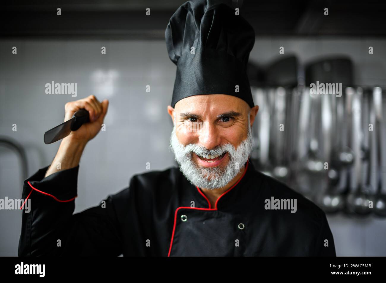 Ein wütender, weißbärtiger Koch in einem schwarzen Mantel, der ein Messer hält, das dich bedroht Stockfoto