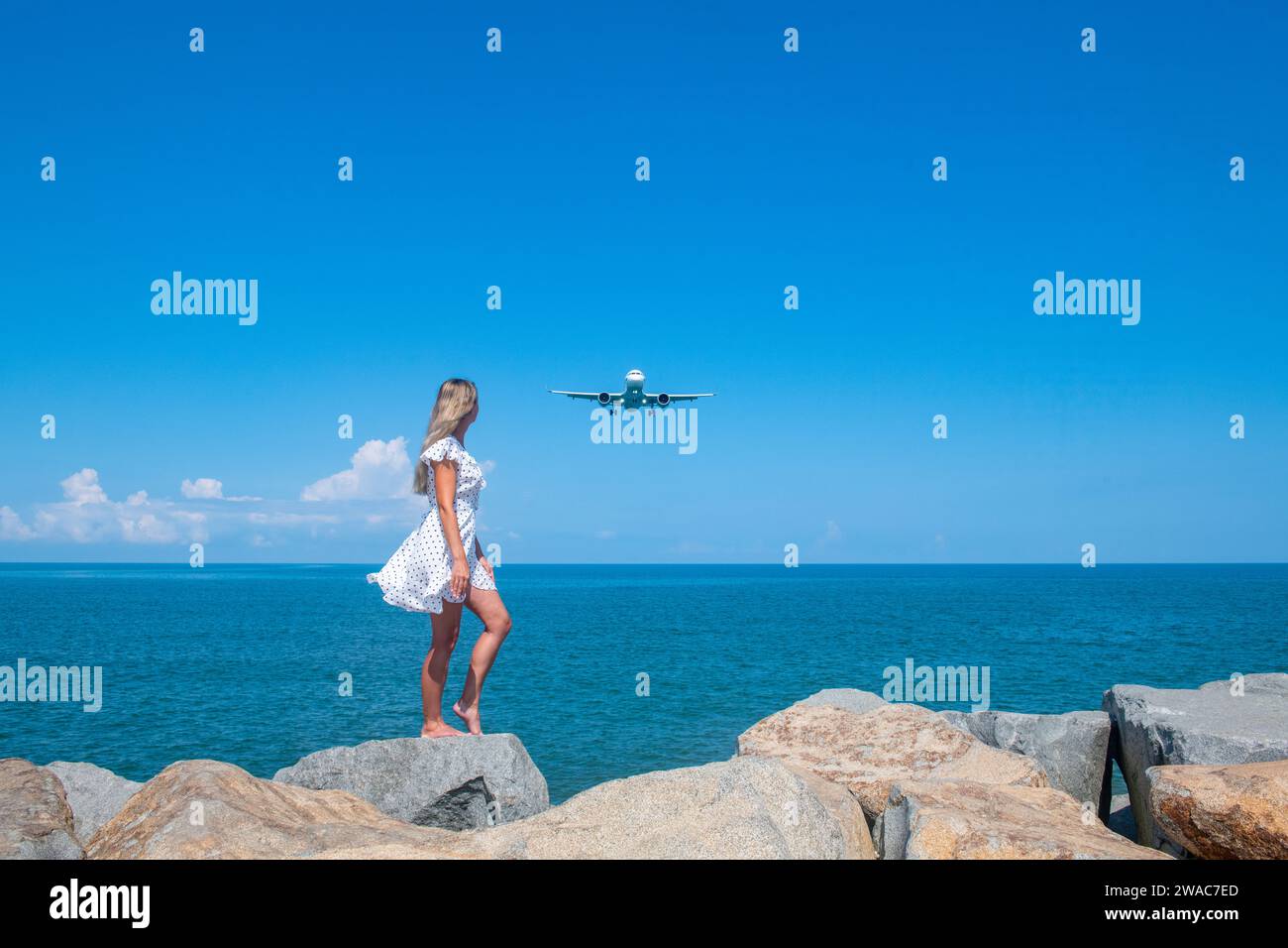 Airborne Encounters: Mädchen in weißem Kleid auf Steinen, Flugzeug über dem Blauen Meer Stockfoto