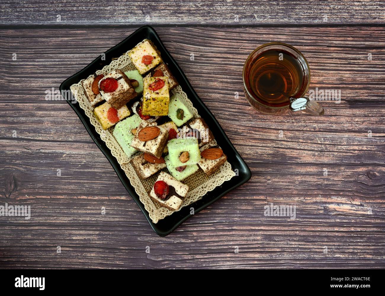 Ein rechteckiger schwarzer Teller mit einer vielfältigen Auswahl orientalischer Süßigkeiten und eine Glasschale heißen Tees mit einem Löffel auf einem Holztisch. Draufsicht, flach. Stockfoto