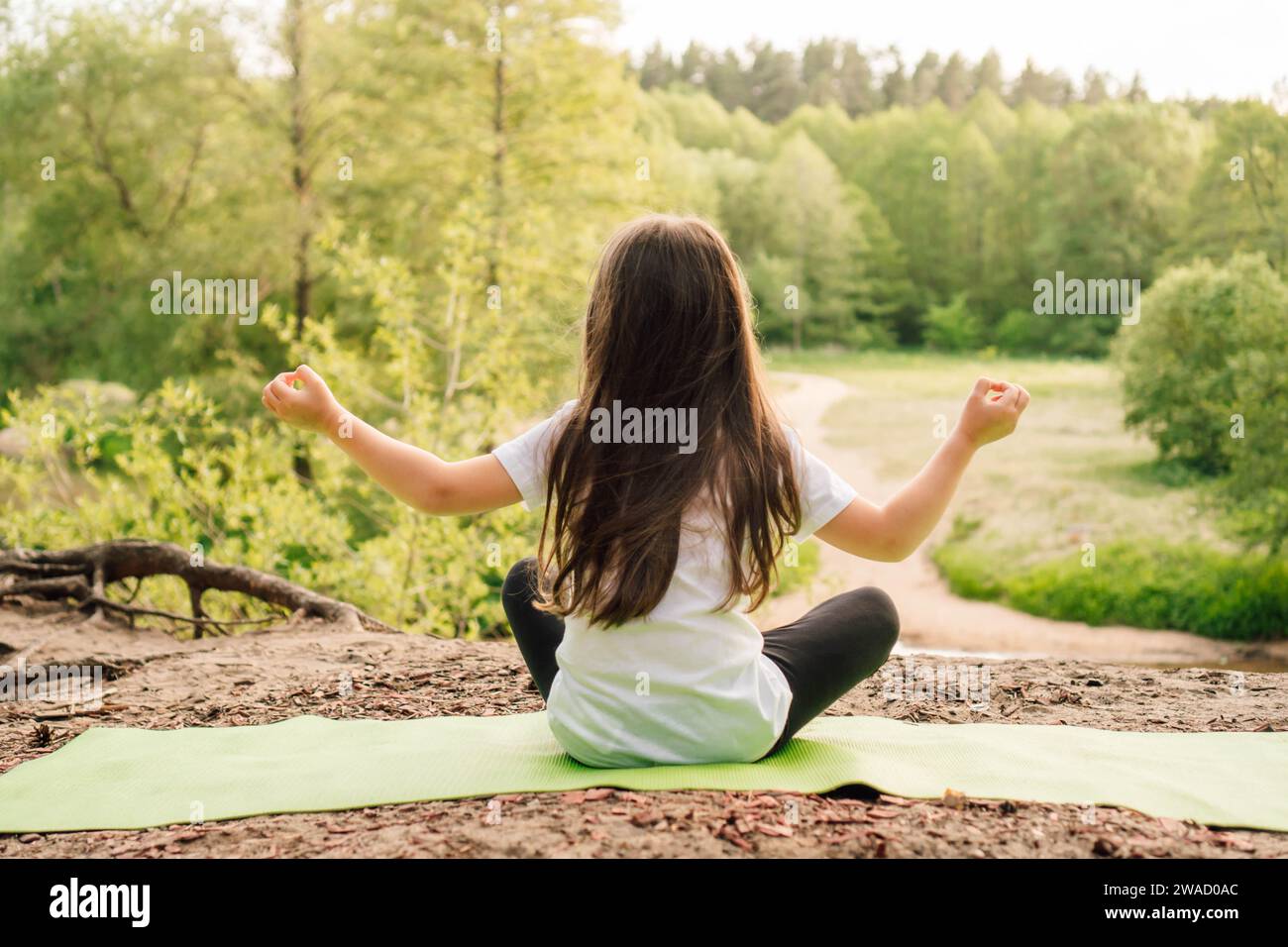 Rückansicht eines kleinen Mädchens mit braunen langen Haaren, das auf grünem, sportlichem Teppich sitzt und vor dem Fluss Yoga praktiziert. Aktive Sportlerin in weißem T-sh Stockfoto