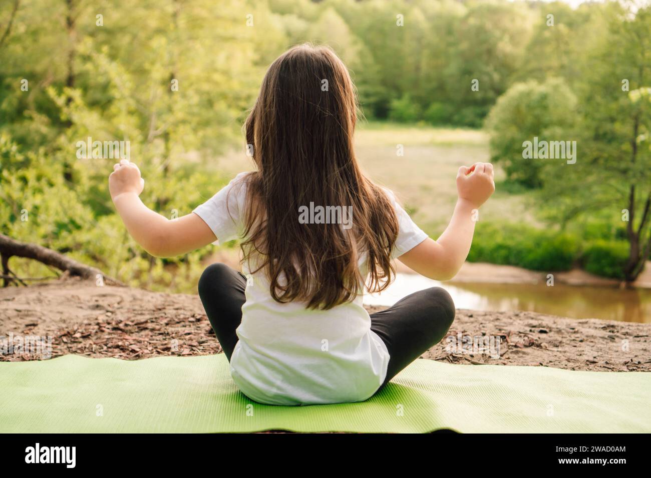 Rückansicht eines kleinen Mädchens mit braunen langen Haaren, das auf grünem, sportlichem Teppich sitzt und vor dem Fluss Yoga praktiziert. Aktive Sportlerin in weißem T-sh Stockfoto