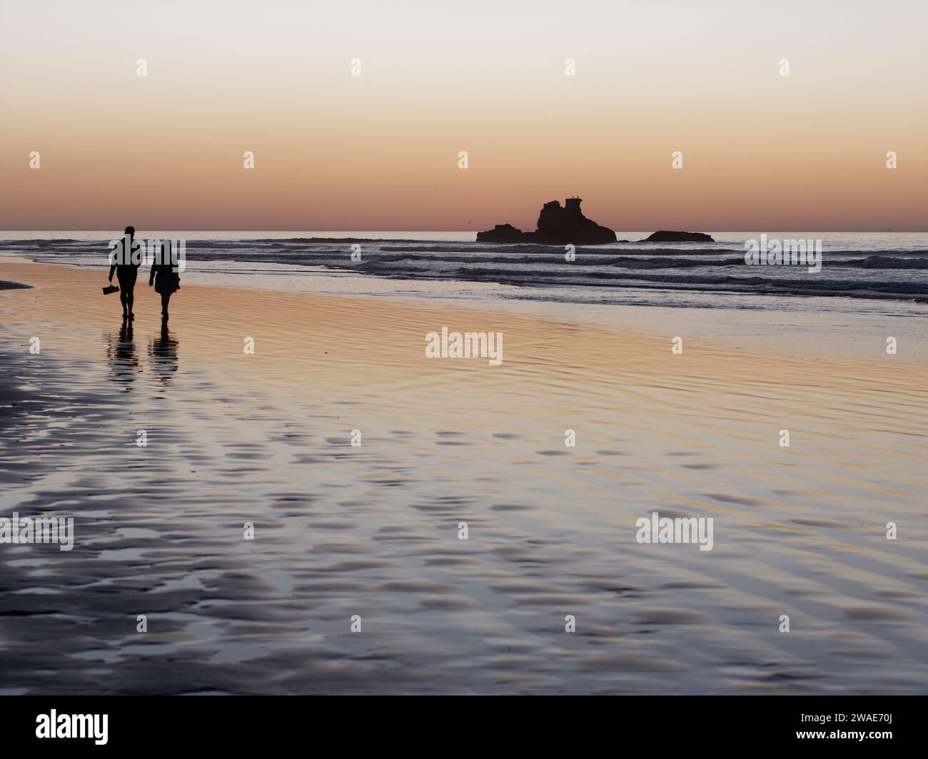 Ein Paar in Silhouette, das bei Sonnenuntergang im seichten Wasser spaziert, mit Felsvorsprüngen dahinter, während der Mann seine Schuhe trägt. Essaouira, Marokko, 3. Januar 2024 Stockfoto