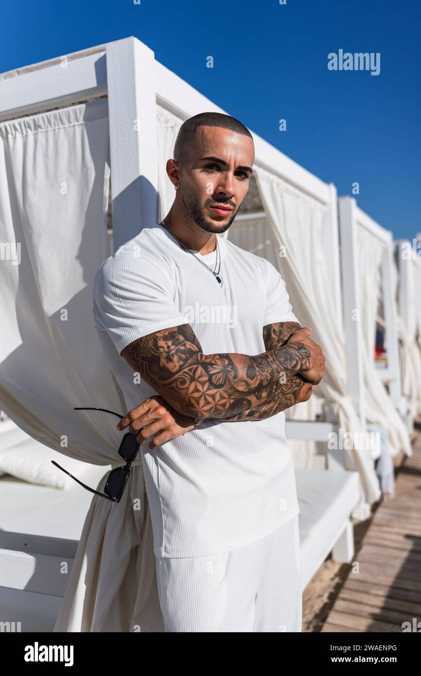Ein bärtiger Mann mit Tätowierungen, der vor einer Reihe weißer Sonnenliegen steht Stockfoto