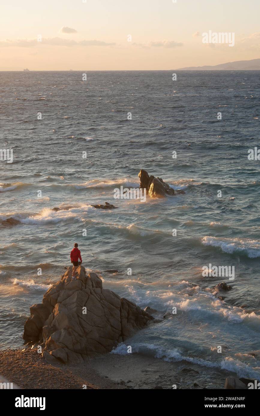 Mann in roter Jacke, der auf Felsen steht und über das wellige Meer blickt, Sonnenuntergang, Casper David Friedrich, Mykonos Stadt, Griechenland, kykladische Insel, ein episches Kinoerlebnis Stockfoto
