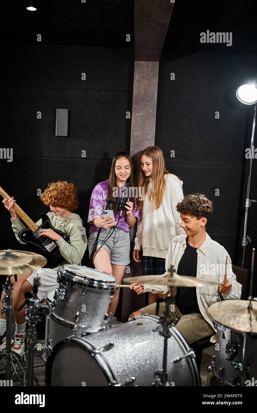 Fröhliche Teenager-Mädchen singen, während Jungs Schlagzeug und Gitarre im Studio spielen Stockfoto