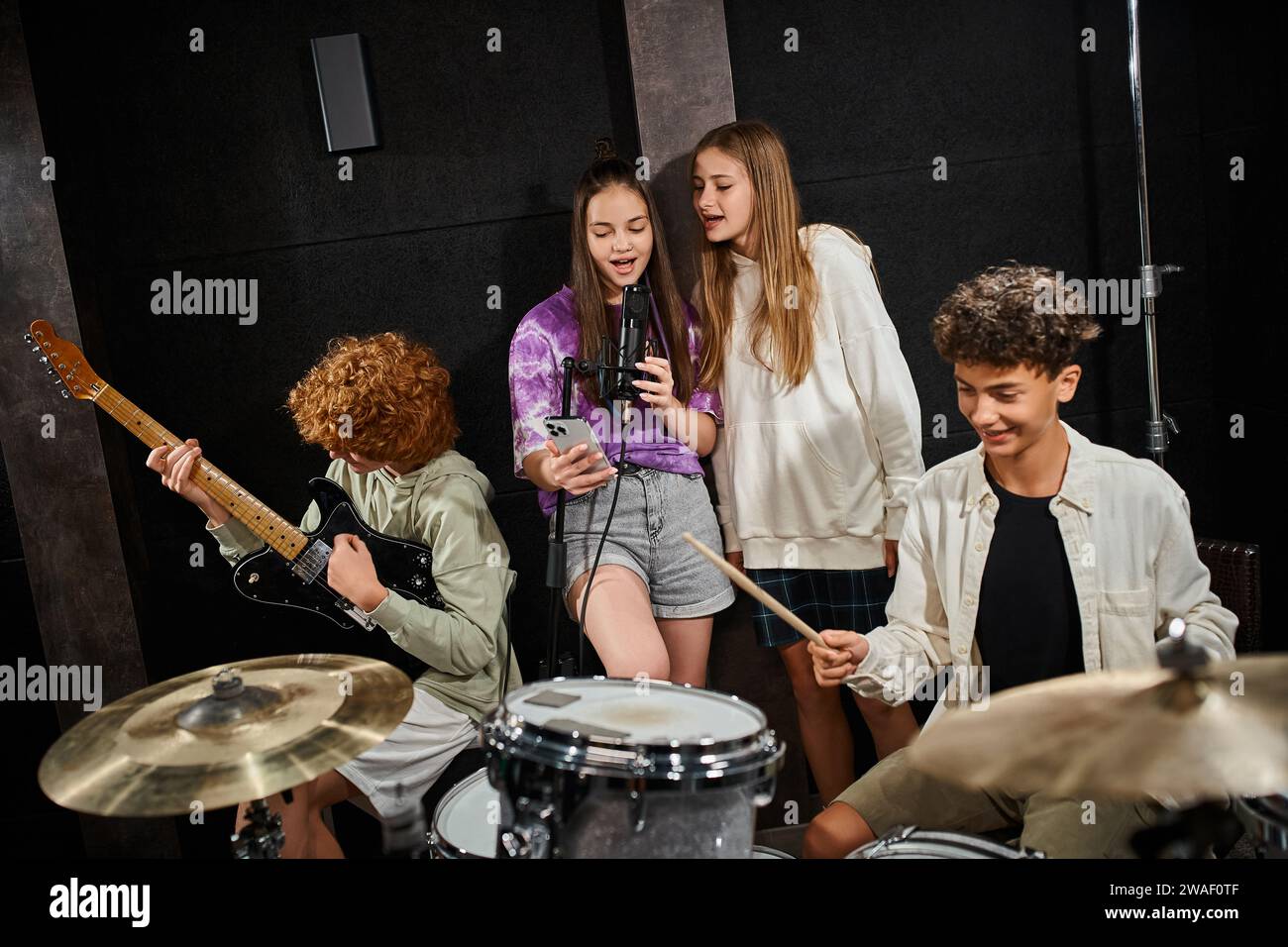 Niedliche Teenager-Jungs in lässigen Kleidern spielen Schlagzeug und Gitarre, während Mädchen Lieder singen, Musikgruppe Stockfoto