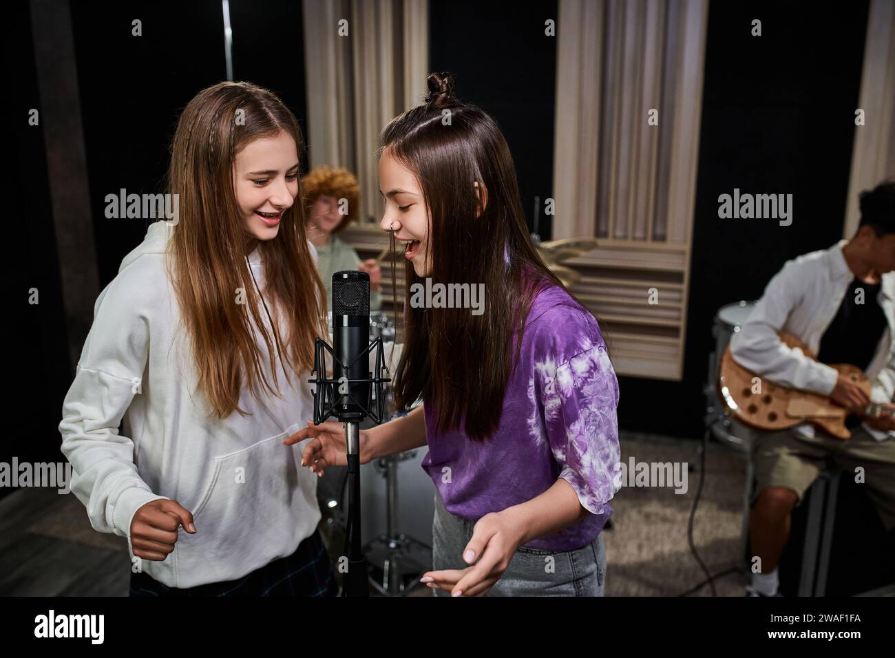 Hübsche Teenager-Mädchen singen ins Mikrofon, während Jungs Schlagzeug und Gitarre spielen, Musikgruppe Stockfoto
