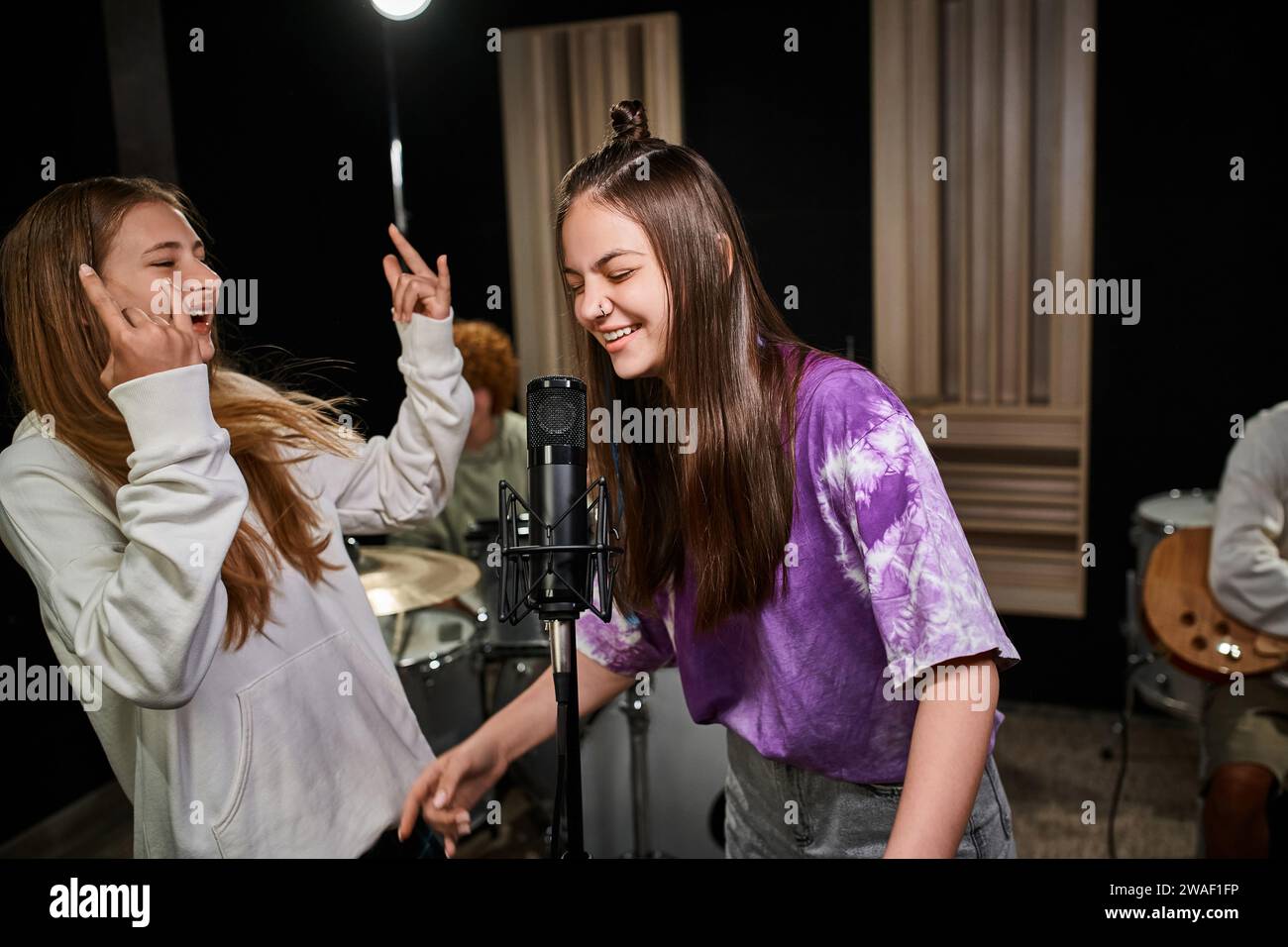 Hübsche Teenager-Mädchen singen und zeigen Rockzeichen, während Jungs Schlagzeug und Gitarre im Studio spielen Stockfoto
