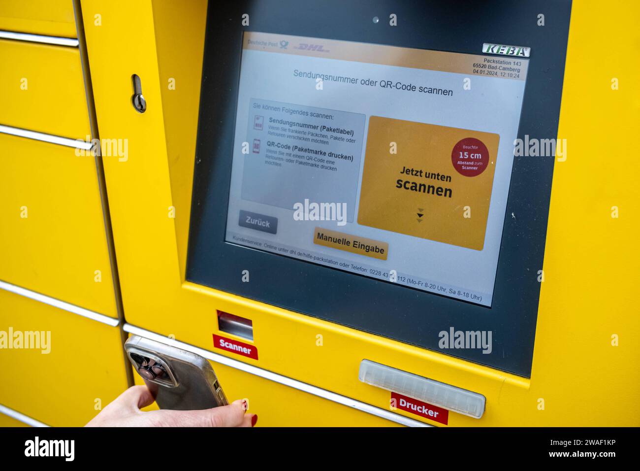 Innovative Paketlogistik - Nutzung der DHL Packstation 04.01.2024, Bad Camberg: Symbolfoto, Illustrationsbild, Symbolbild, Illustrationsfoto das Bild zeigt eine Frau im Anschnitt, die mit ihrem Smartphone über den Touchscreen einer DHL Paketstation interagiert, um ein Paket einzureichen. Sie ist konzentriert auf die digitale Schnittstelle fokussiert, während Ihre Hand das Mobiltelefon hält und die entsprechenden Eingaben auf dem Bildschirm der Paketstation tätigt um das Paket einlegen. Die Packstation, ein Paketautomatensystem von DHL, bietet seit 2001 in über 12,500 Standorten deutschlandwe Stockfoto