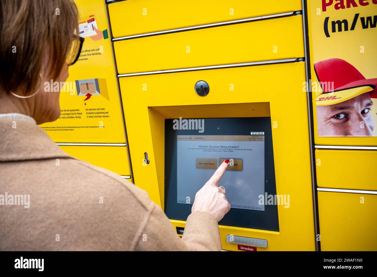 Innovative Paketlogistik - Nutzung der DHL Packstation 04.01.2024, Bad Camberg: Symbolfoto, Illustrationsbild, Symbolbild, Illustrationsfoto das Bild zeigt eine Frau im Anschnitt, die mit ihrem Smartphone über den Touchscreen einer DHL Paketstation interagiert, um ein Paket einzureichen. Sie ist konzentriert auf die digitale Schnittstelle fokussiert, während Ihre Hand das Mobiltelefon hält und die entsprechenden Eingaben auf dem Bildschirm der Paketstation tätigt um das Paket einlegen. Die Packstation, ein Paketautomatensystem von DHL, bietet seit 2001 in über 12,500 Standorten deutschlandwe Stockfoto