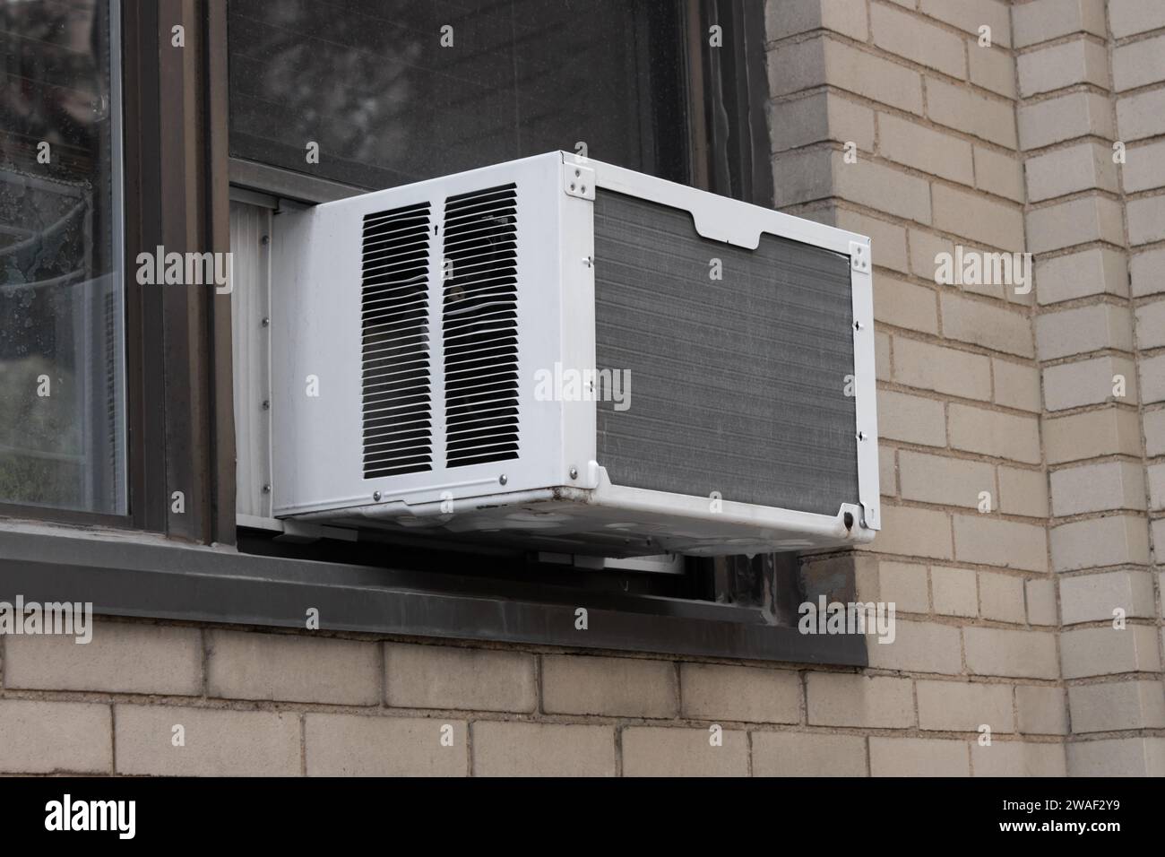 Außenansicht der Klimaanlagen-Fenstereinheit, die von der Fensterbank eines beigefarbenen Ziegelgebäudes aus extrudiert wird Stockfoto