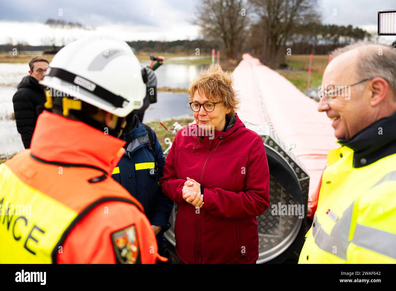 04. Januar 2024, Niedersachsen, Winsen (aller): Daniela Behrens (SPD, 2. V. R.), niedersächsische Innenministerin und Sportministerin, spricht bei ihrem Besuch bei den französischen Zivilschutzkräften mit Oberstleutnant-Oberst Benoit Pinaud (l), Teamleiterin der französischen Zivilschutzkräfte. Neben ihm steht Dieter Rohrberg (r), Landesfeuerwehrdirektor Niedersachsens. Im Hintergrund sehen Sie einen Teil eines mobilen Deiches, der mit Wasser aus der aller gefüllt ist. Ein Team des französischen Zivilschutzes wird bei Thören in der Gemeinde Winsen (aller) einen mobilen Deich bauen. Das ist ein Europa-Wid Stockfoto